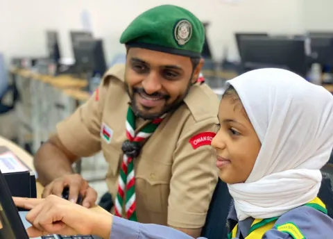 An Omani Scout Leader sits at a computer with a Scout girl during JOTA-JOTI 2021