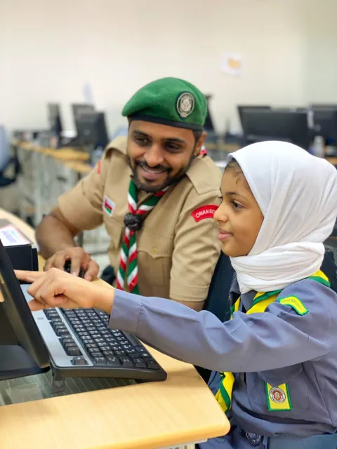 An Omani Scout Leader sits at a computer with a Scout girl during JOTA-JOTI 2021