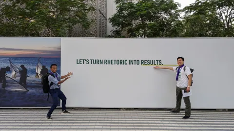 WOSM Youth Representatives Kek and Lucas pose at COP28 in front of a wall which reads "Let's turn rhetoric into action"
