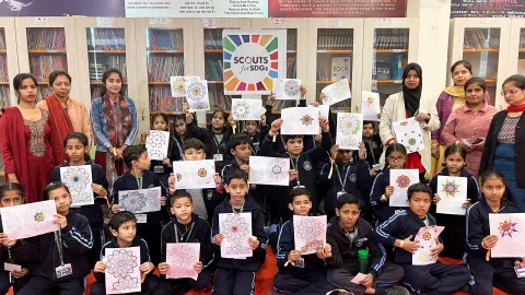 A group of young students at Vrinda International School in New Delhi, holding up their colorful mandala art. They're gathered in the library with 'Scouts for SDGs' prominently displayed, showcasing their participation in World Scouting’s Mental Health Challenge.
